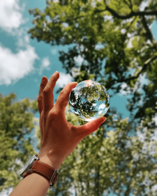 Uma mão segura uma bola de video, ela reflete a paisagem de árvores e céu a sua volta, formando uma espécie de mini planeta.